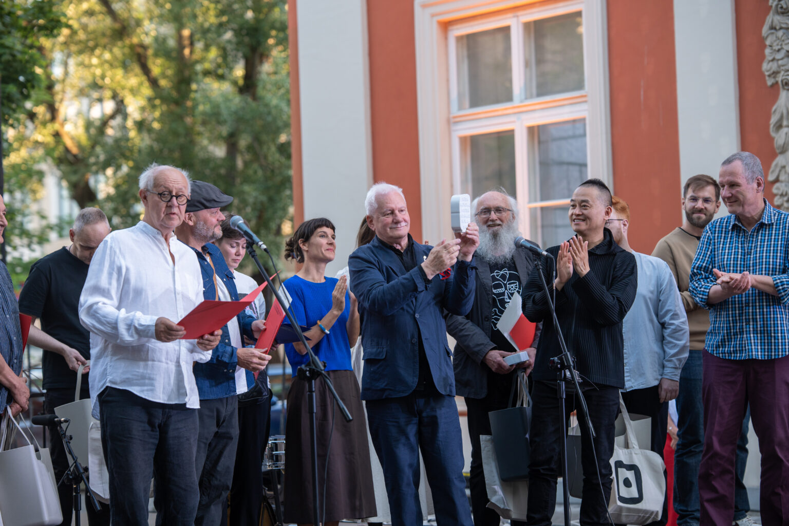 Mężczyźni i kobieta w różnym wieku, stojący na scenie. Jeden mężczyzna w białej koszuli trzyma czerwone dokumenty, a inni biją brawo.