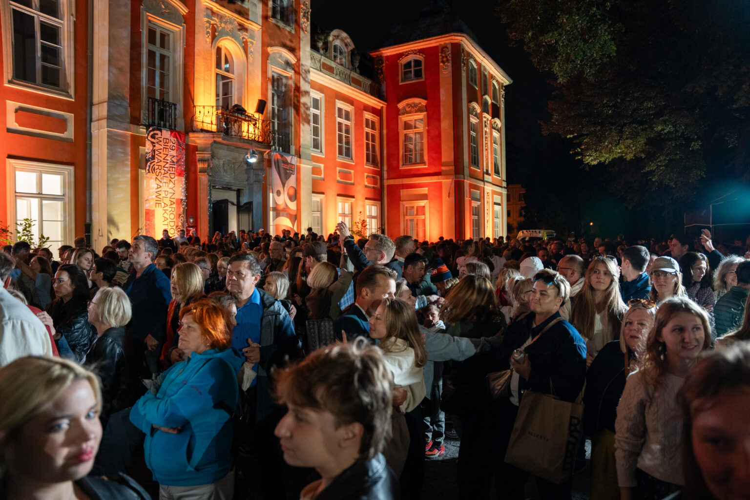 Tłum ludzi zebrał się na zewnątrz, aby wziąć udział w nocnej imprezie. Budynek w tle jest podświetlony.
