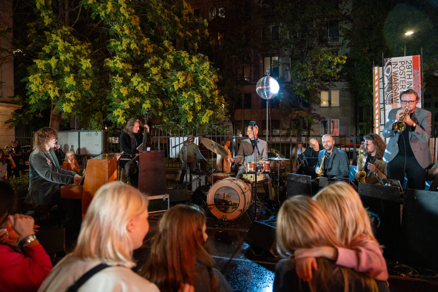 Ludzie oglądają koncert na świeżym powietrzu. Na scenie gra zespół jazzowy.