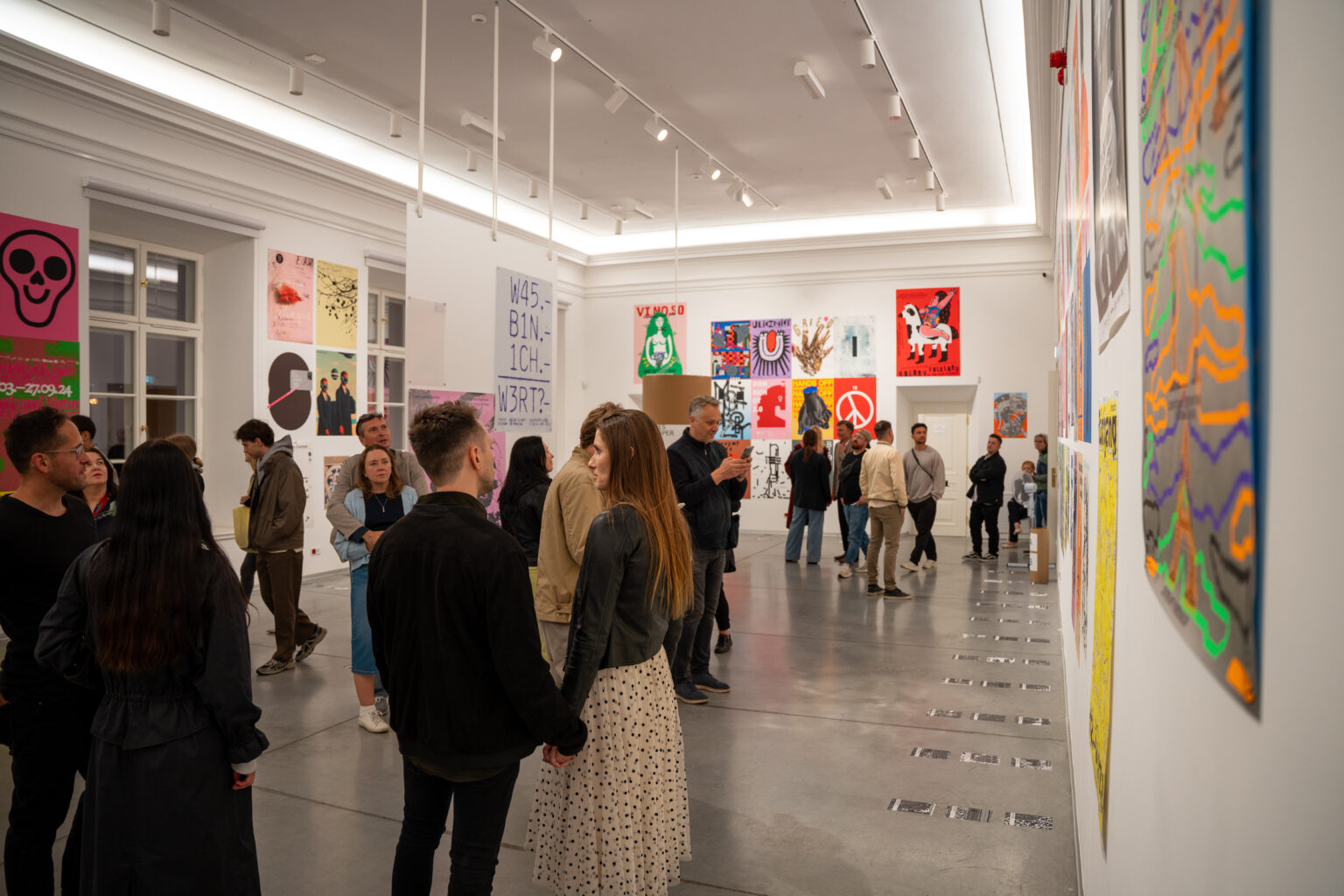 Ludzie oglądają wystawę sztuki. W tle widać plakaty w różnych stylach i kolorach.