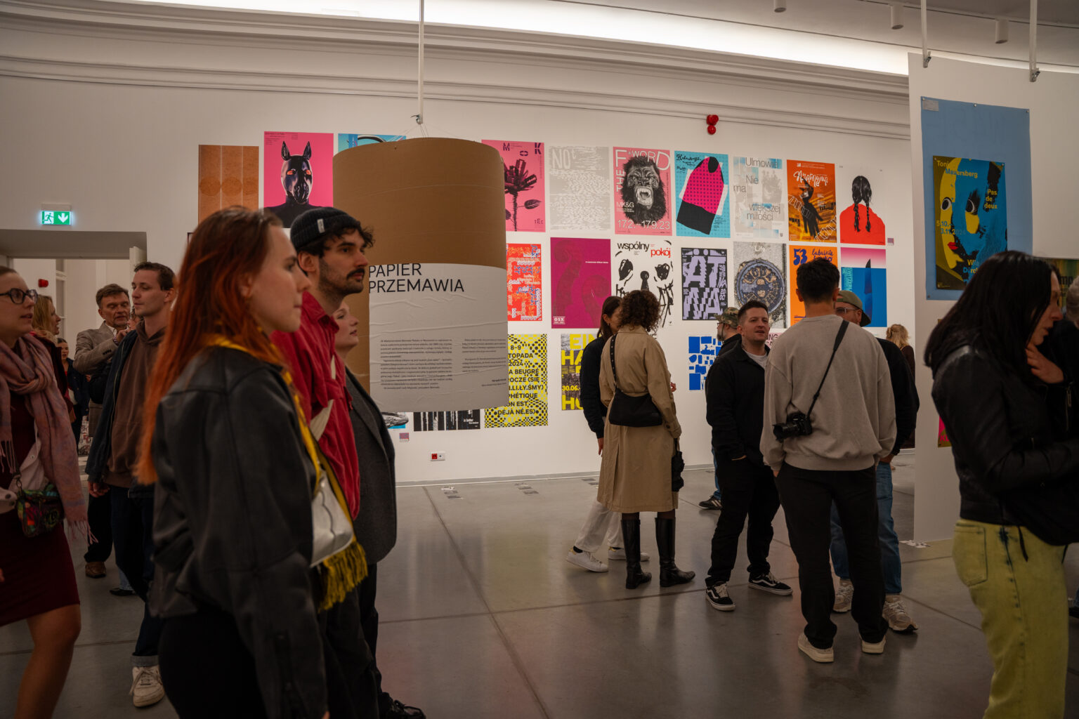 Ludzie oglądają wystawę sztuki. W tle widać plakaty w różnych stylach i kolorach.