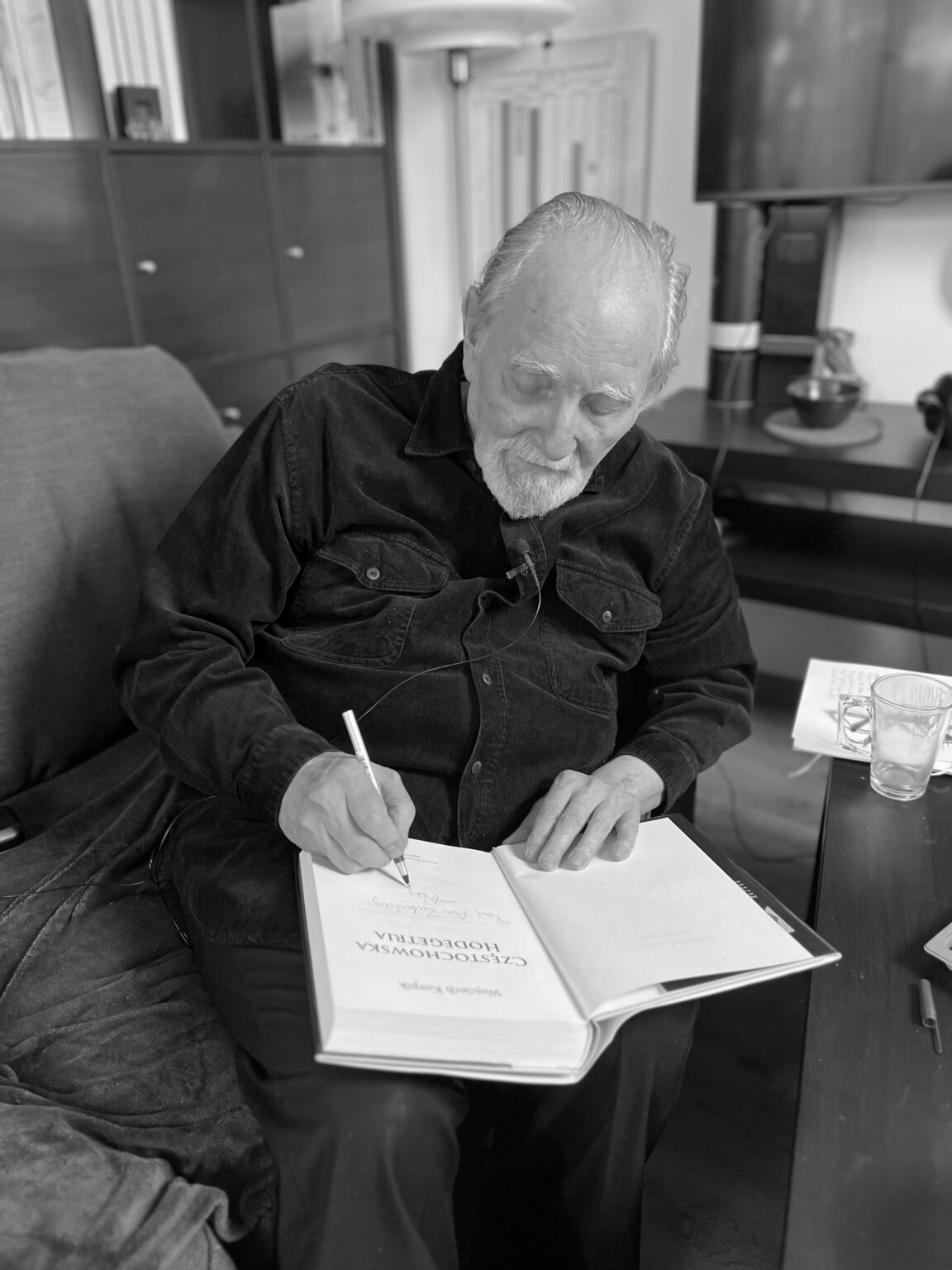 Czarno-białe zdjęcie siedzącego mężczyzny w ciemnej koszuli, podpisującego książkę.
