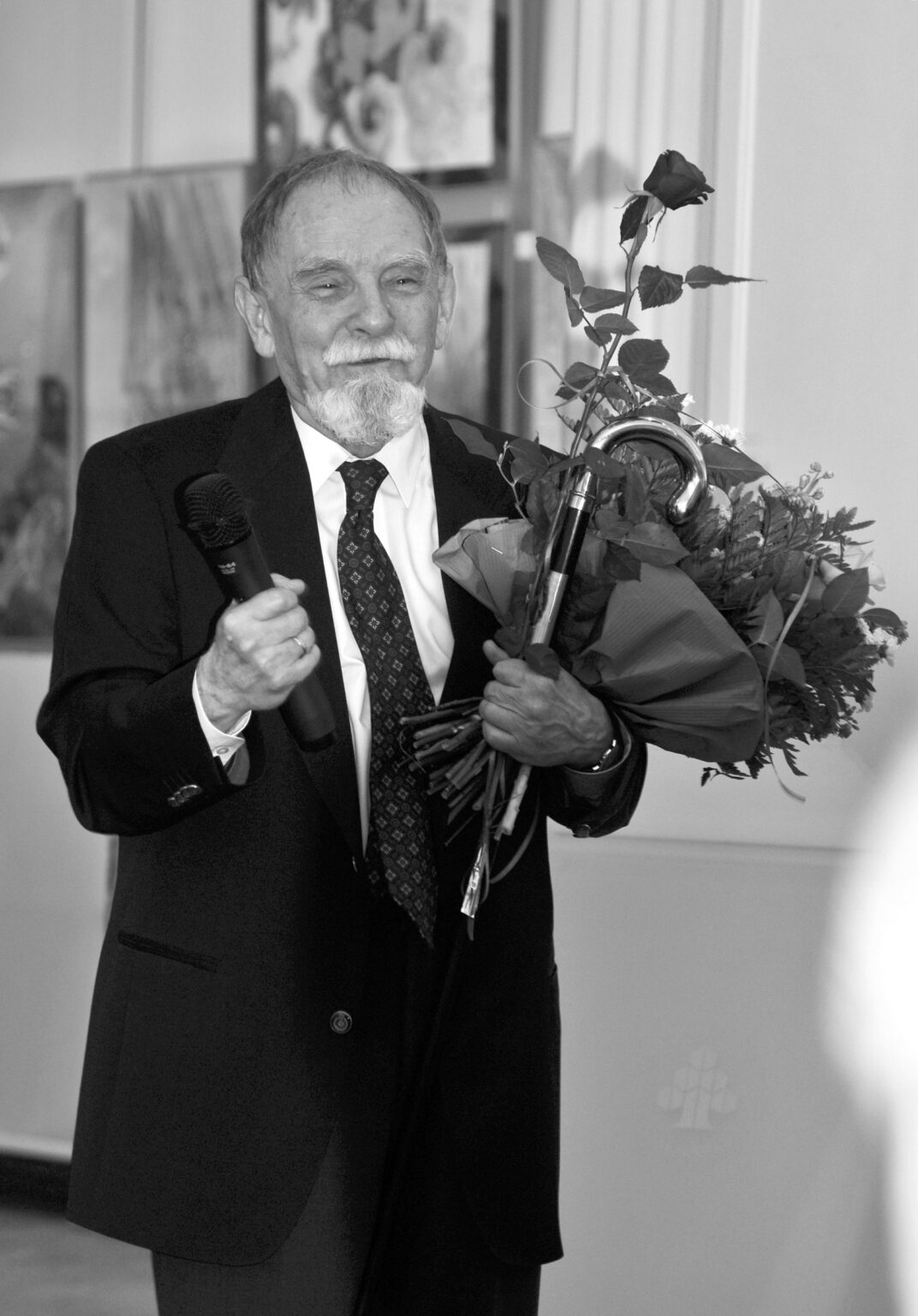 Czarno-białe zdjęcie starszego mężczyzny w garniturze z brodą i wąsami, trzymającego mikrofon i bukiet kwiatów.