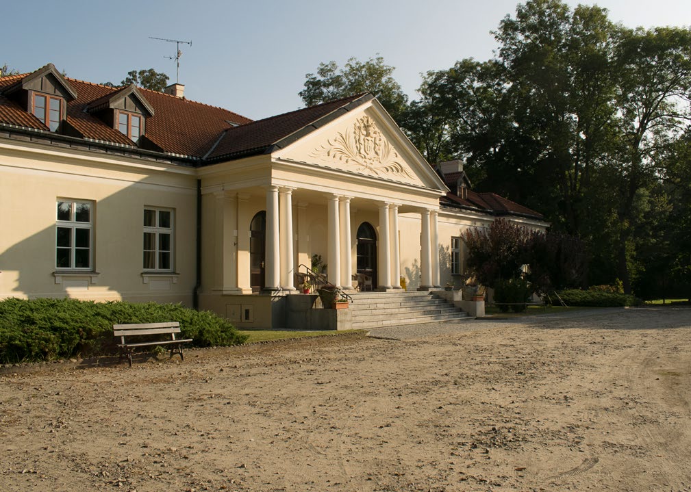 Biały, klasycystyczny dwór z czerwonym dachem i kolumnowym portykiem, otoczony zielenią, przed nim ławka i piaszczysty podjazd