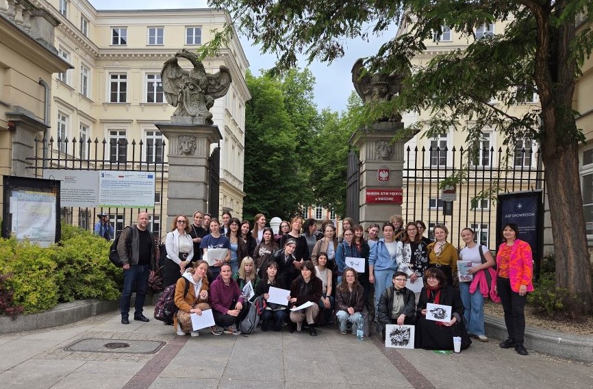 Grupa ludzi pozuje do zdjęcia przed bramą dużego, starego budynku. Na bramie widoczne są rzeźby orłów.