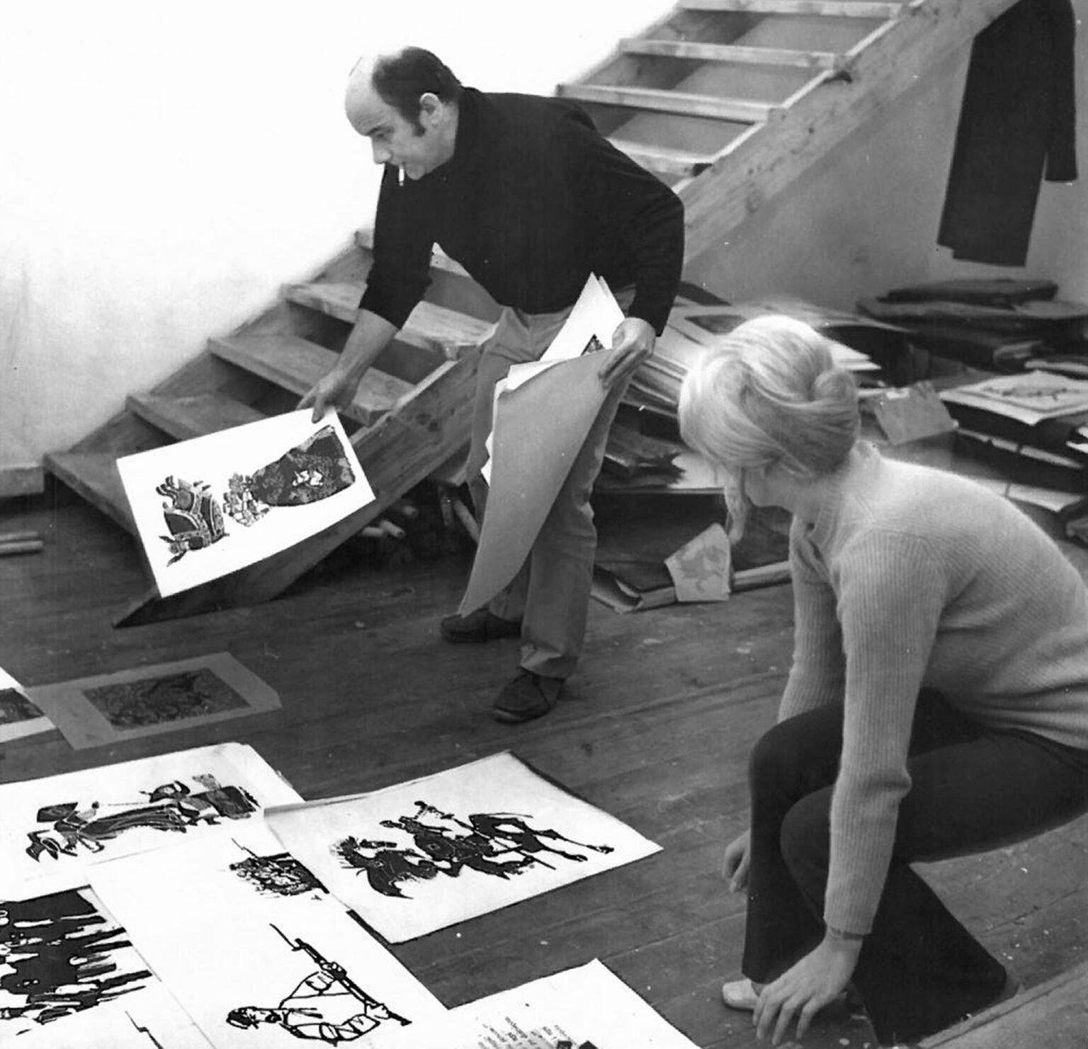 Dwoje artystów ogląda grafiki rozłożone na podłodze. Mężczyzna schylony trzyma jeden z obrazów, kobieta kuca obok.