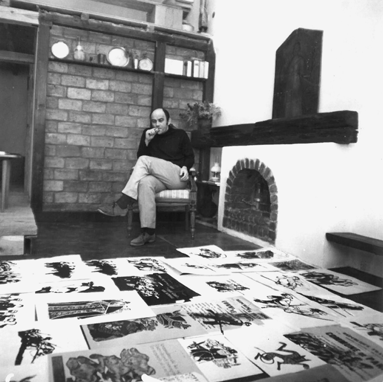 Mężczyzna siedzi na krześle, zamyślony, obok kominka. Na podłodze i stole leżą rozłożone grafiki.