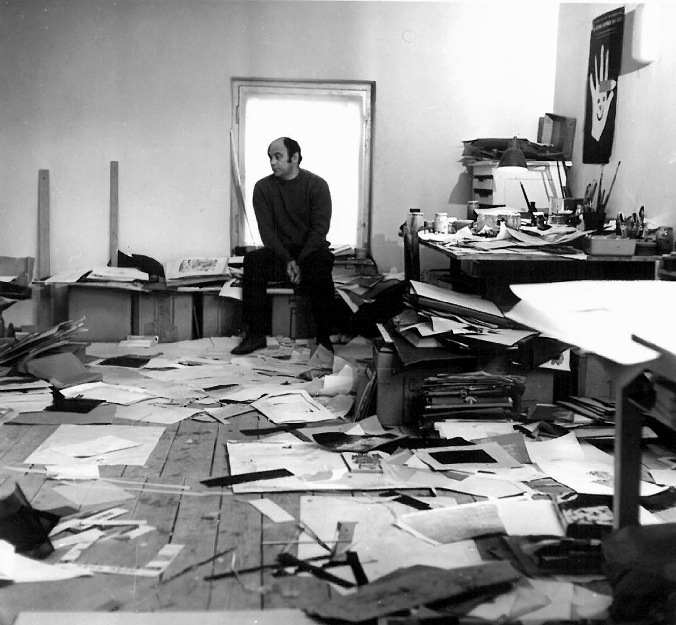 Artysta siedzi w pracowni artystycznej na tle okna. Na podłodze i biurkach panuje artystyczny nieład.