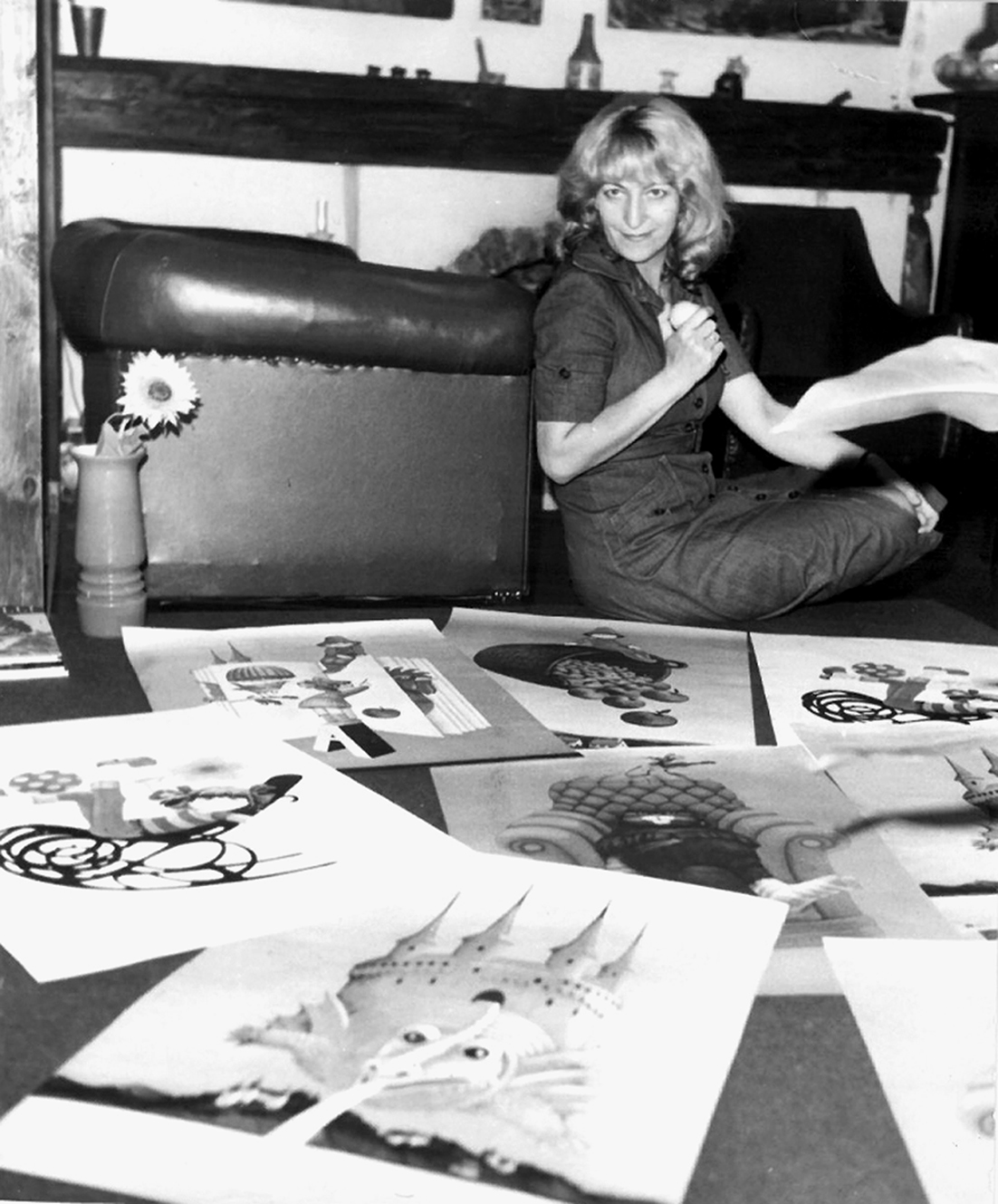 Kobieta siedzi na podłodze, otoczona grafikami. Uśmiecha się, trzymając w ręku jeden z obrazów, a drugi rozkłada.