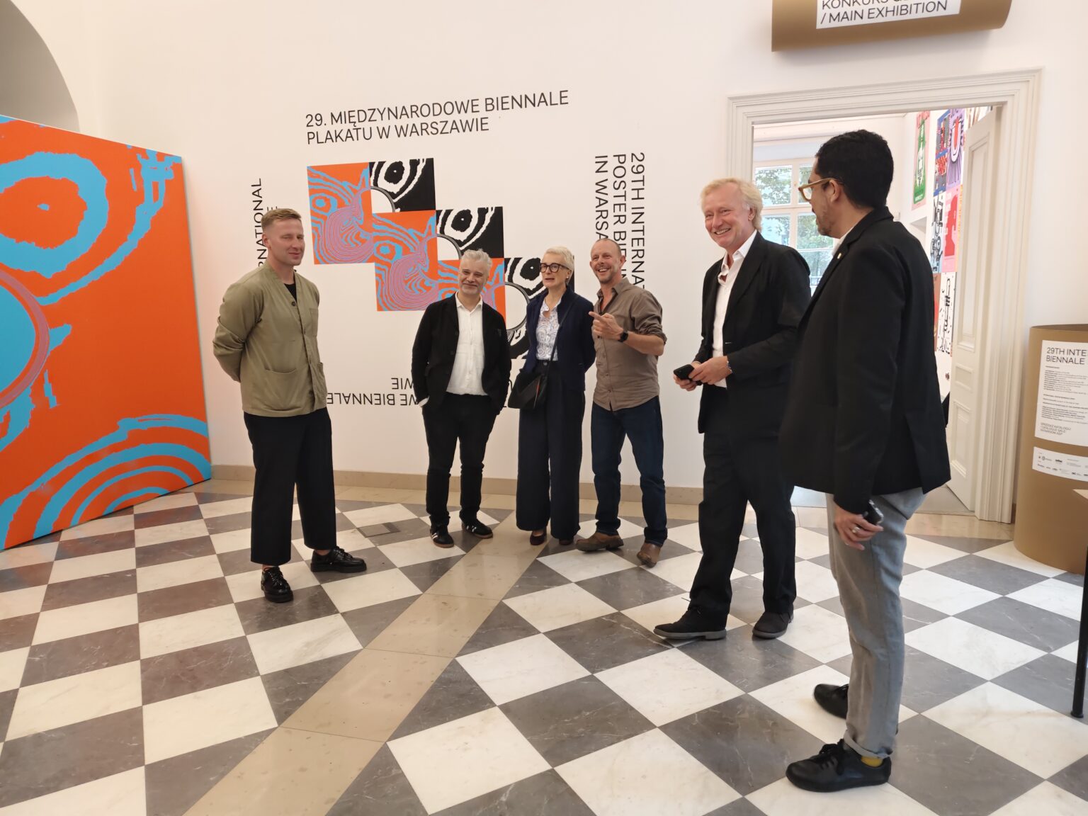 Grupa osób rozmawia w galerii sztuki, na tle ściany z plakatem promującym 29. Międzynarodowe Biennale Plakatu w Warszawie.