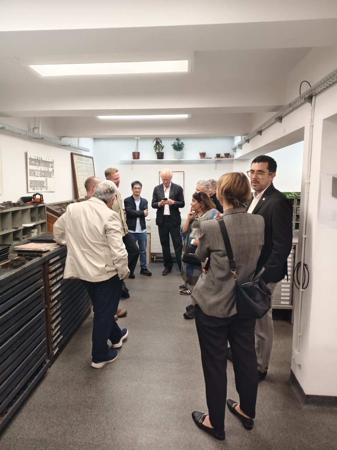 Grupa osób stoi w wąskim pomieszczeniu z półkami wypełnionymi szufladami. Pośrodku dyskutują, a jeden z mężczyzn używa telefonu.