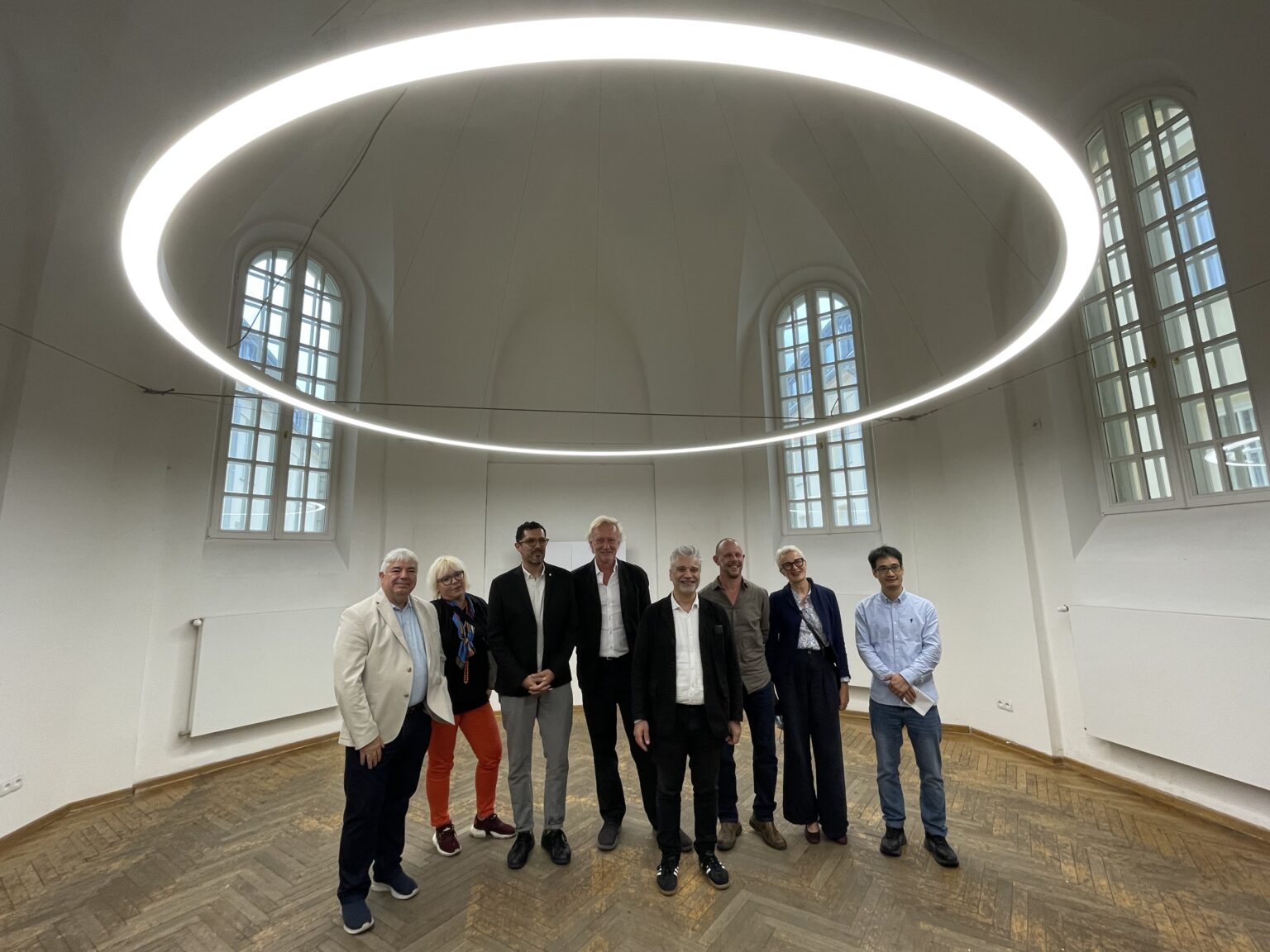 Duża, podświetlana obręcz zwisa z sufitu. Pod nią stoi grupa ośmiu osób w przestronnym, pustym pomieszczeniu z drewnianą podłogą.