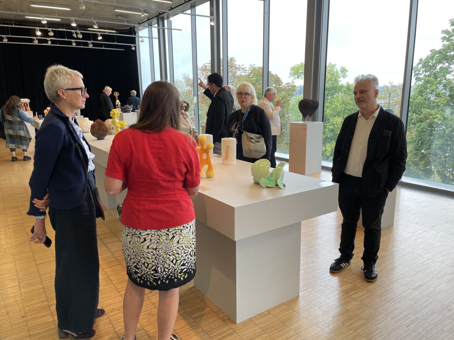 Kilka osób ogląda eksponaty na białych, minimalistycznych stołach w galerii sztuki, oświetlonej naturalnym światłem z dużych okien.