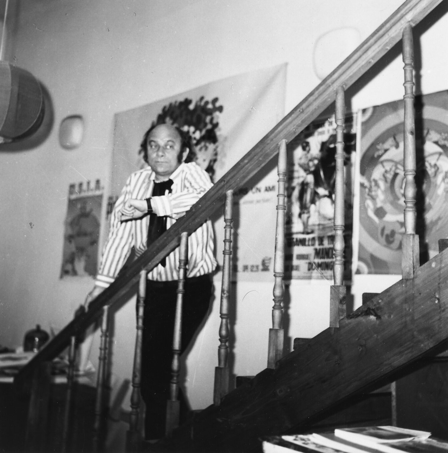 Mężczyzna stoi na schodach, opierając się o balustradę. Za nim widać plakaty. Ubrany jest w koszulę w paski i ciemne spodnie.