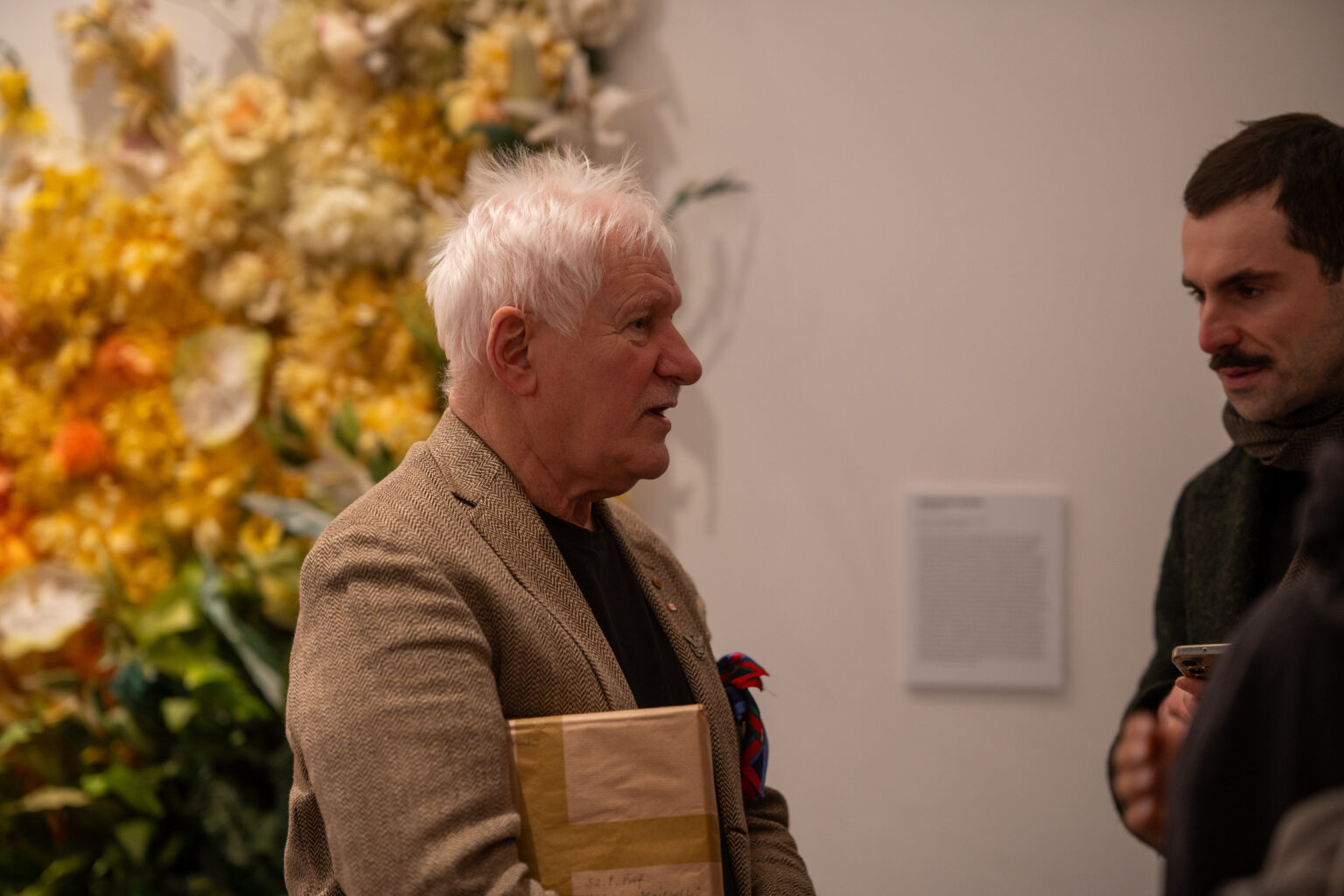 mężczyzna z siwymi włosami w beżowej marynarce rozmawia z drugą osobą w galerii. W tle widać obfitą instalację z żółtych i jasnych kwiatów oraz białą ścianę z opisem.