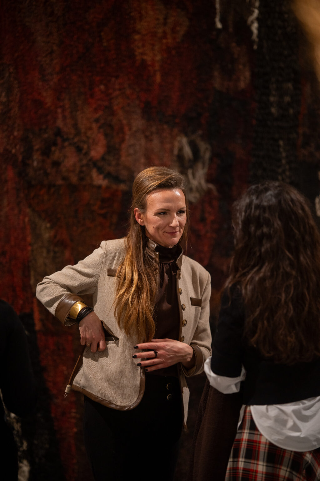 Kobieta w beżowym żakiecie i brązowym golfie uśmiecha się podczas rozmowy w galerii. W tle widać fragment monumentalnej, ciemnoczerwonej tkaniny artystycznej.