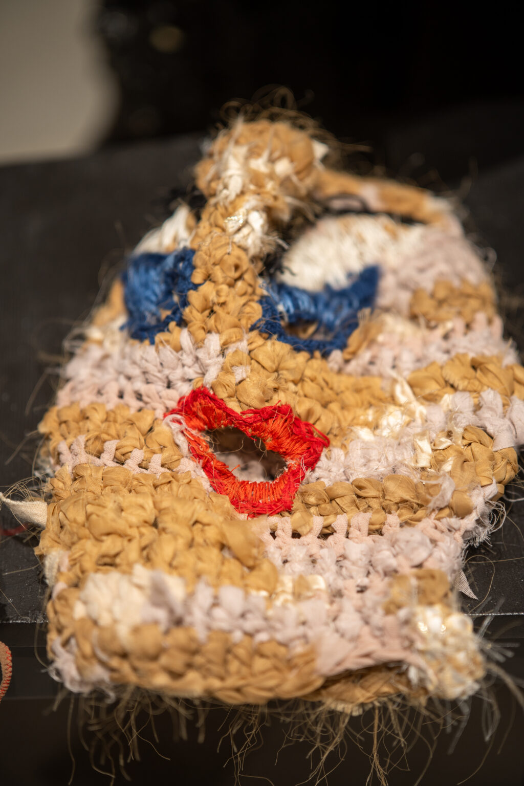 Zbliżenie na maskę wykonaną z grubych, plecionych sznurków i tkanin. Obiekt ma wyraźnie zaznaczone czerwone usta oraz niebieskie akcenty wokół otworów na oczy.