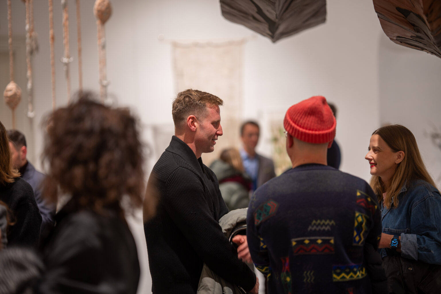 Na pierwszym planie mężczyzna w czarnym swetrze i osoba w czerwonej czapce. W tle widać wiszące instalacje artystyczne i inne osoby.