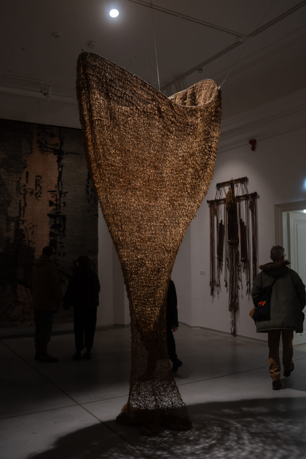 Wysoka, ażurowa rzeźba z brązowej siatki zwężająca się ku dołowi, przypominająca odwrócony stożek. W słabo oświetlonej galerii widać zwiedzających i inne instalacje.