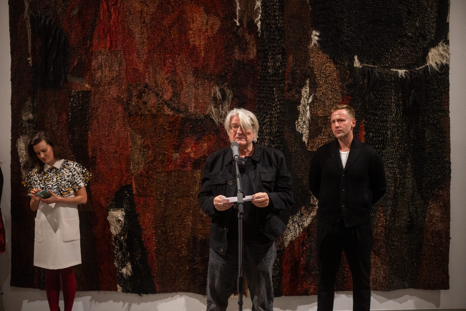 Mężczyzna przemawia do mikrofonu na tle ogromnej, ciemnej tkaniny o bogatej fakturze. Po jego bokach stoją dwie inne osoby, a całość odbywa się w przestrzeni wystawowej.
