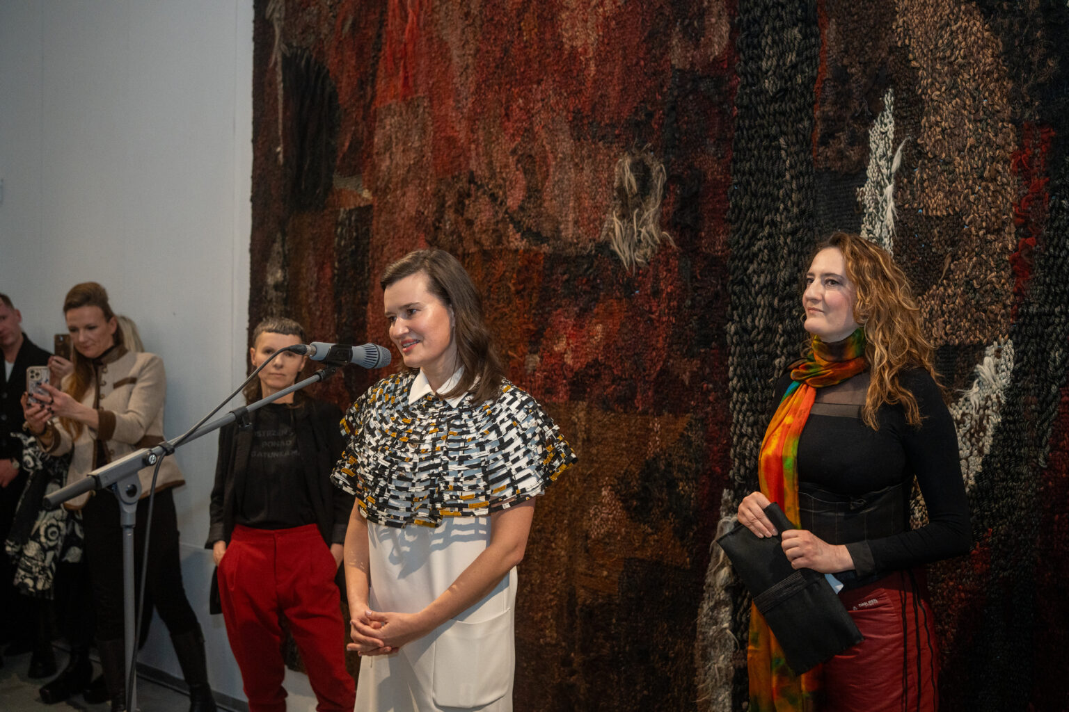 Uśmiechnięta kobieta w białej sukni z ozdobnym kołnierzem przemawia do mikrofonu. Obok niej stoi inna kobieta w kolorowym szalu na tle monumentalnej, ciemnej instalacji z tkanin.