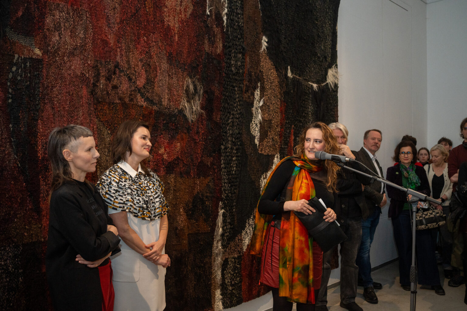 Kobieta w kolorowym szalu przemawia do mikrofonu, trzymając notatki. Obok stoją dwie inne kobiety; w tle widać wiellką tkaninę oraz grupę słuchających gości.