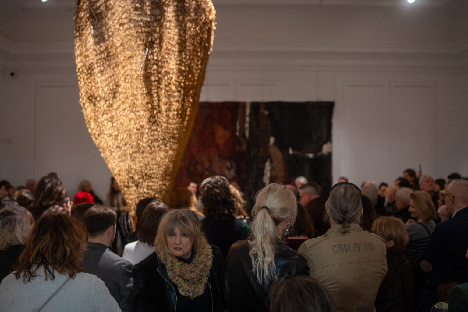 Tłum ludzi zgromadzony w galerii podczas wernisażu. Nad głowami zwiedzających góruje wielka, ażurowa rzeźba z brązowej siatki, a w tle widać monumentalną, ciemną tkaninę.