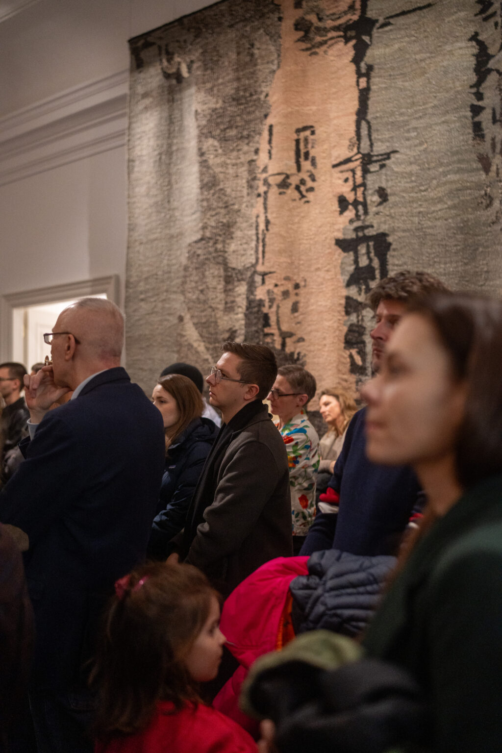 Grupa osób w różnym wieku ogląda wystawę w galerii. W tle widać fragment jasnej tkaniny z czarnymi, pionowymi wzorami przypominającymi abstrakcyjne figury.