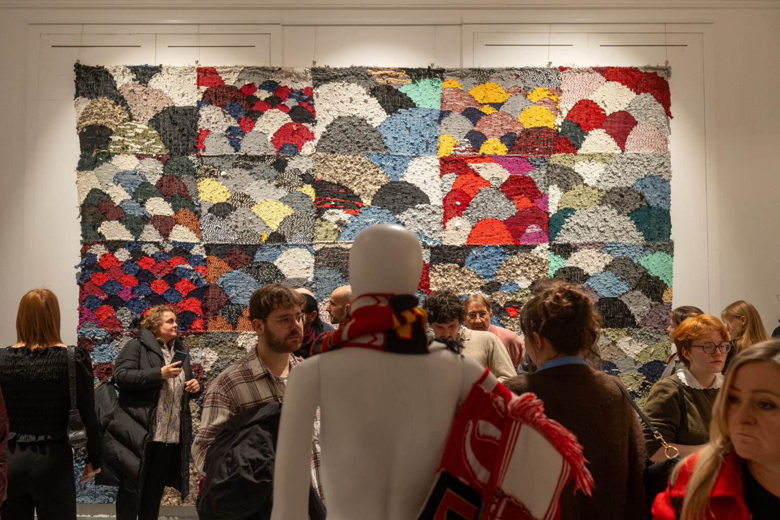 Tłum ludzi przed ogromną, kolorową instalacją z tkanin w galerii. Na pierwszym planie biały manekin tyłem, owinięty czerwono-białym szalikiem;.