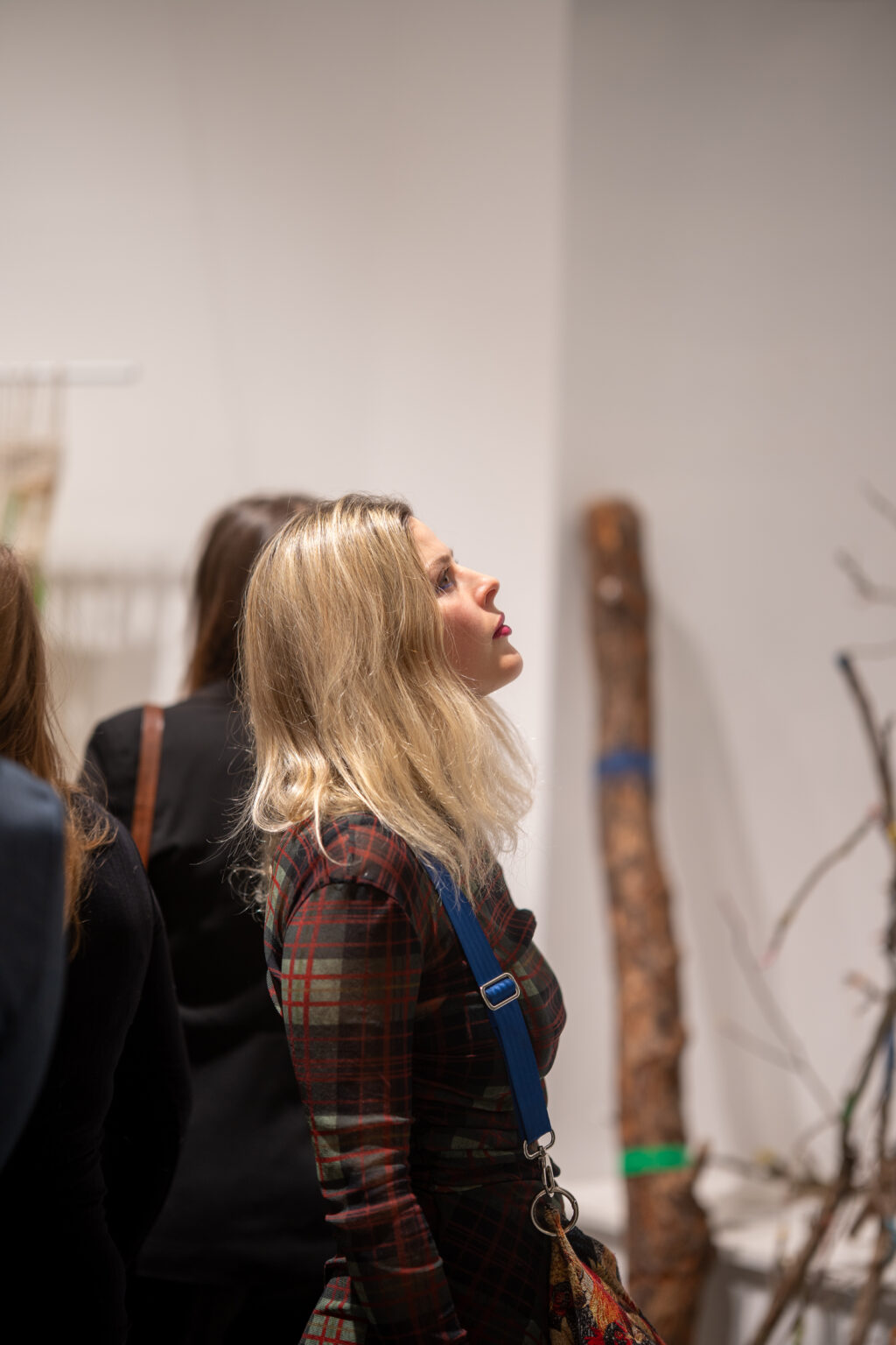 Profil blondynki w ciemnej sukience w kratę, patrzącej w górę na ekspozycję. W tle widać jasną ścianę galerii i fragment drewnianej rzeźby lub instalacji.