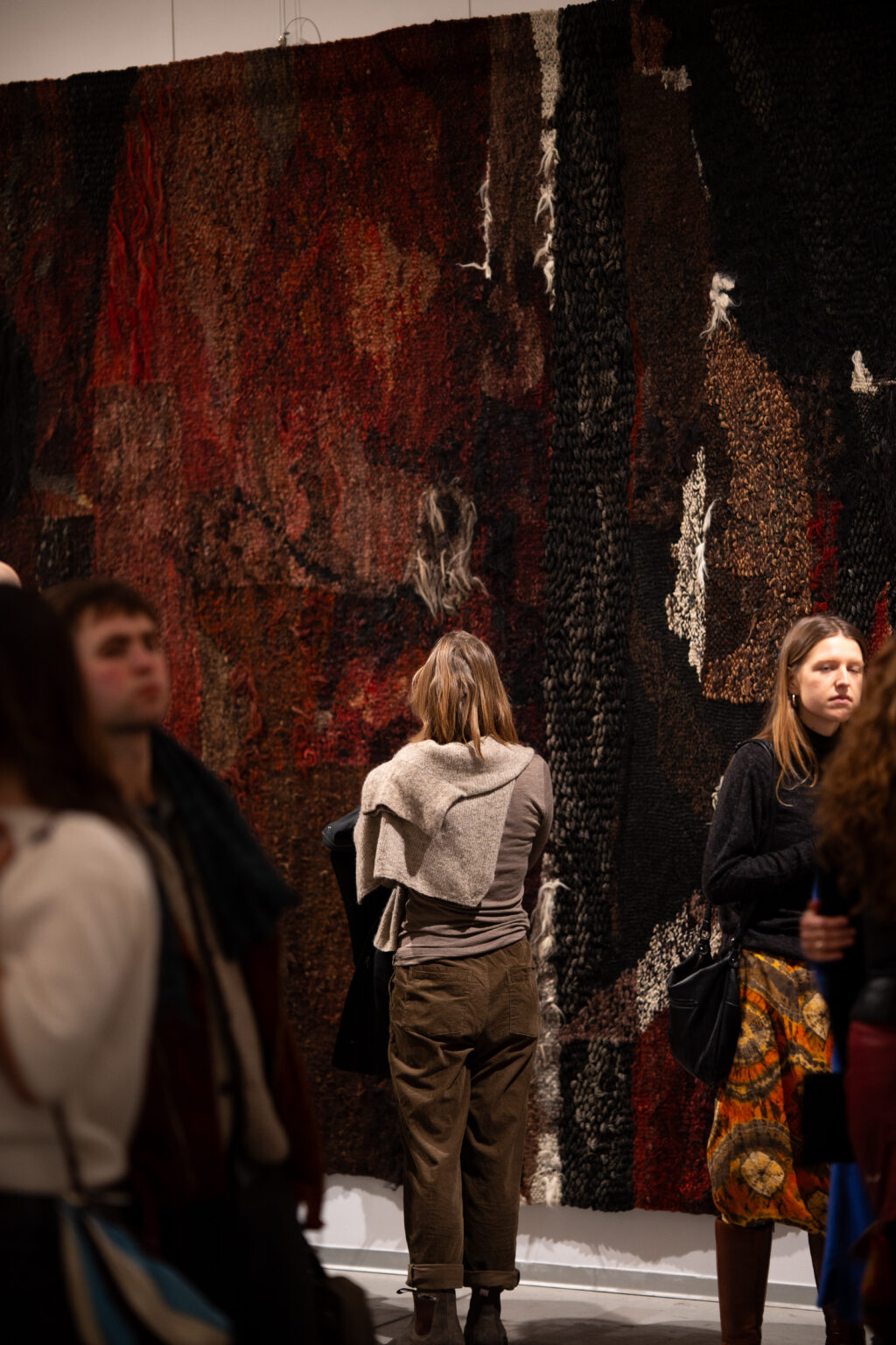 obieta stoi przed ciemną tkaniną o bogatej teksturze w odcieniach brązu, czerni i głębokiej czerwieni. Wokół niej widać fragmenty sylwetek innych osób zwiedzających galerię.