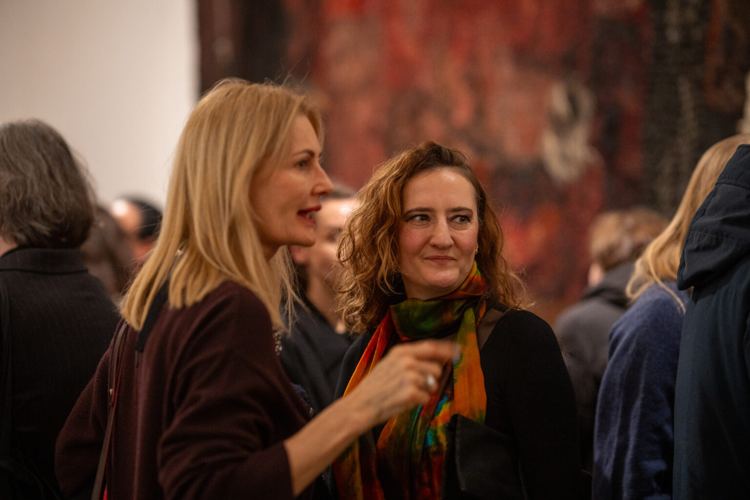 Dwie kobiety rozmawiają w tłumie gości w galerii. Jedna ma na sobie kolorowy, wzorzysty szal; w rozmytym tle widać fragment czerwono-brązowej instalacji.