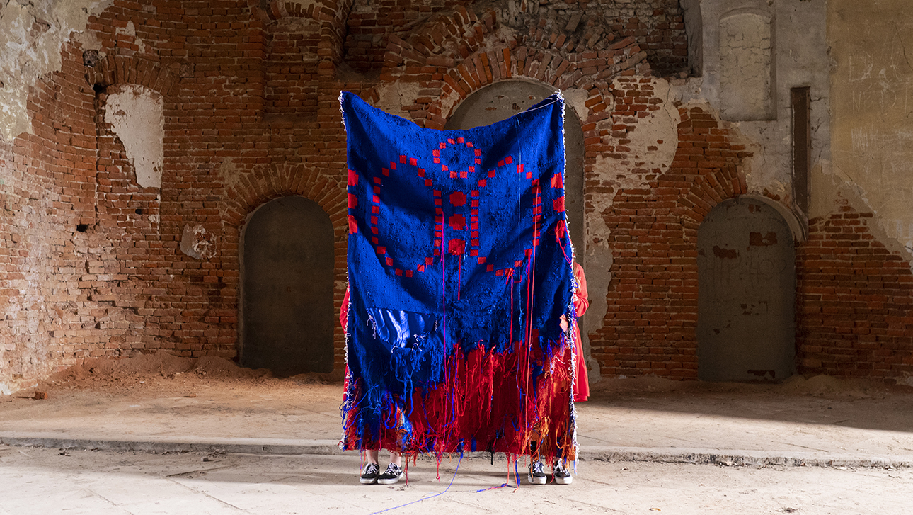 Dwie osoby trzymają dużą, niebiesko-czerwoną tkaninę o nieregularnych brzegach w surowym, ceglanym wnętrzu z łukami. Materiał zdobią czerwone, geometryczne wzory.