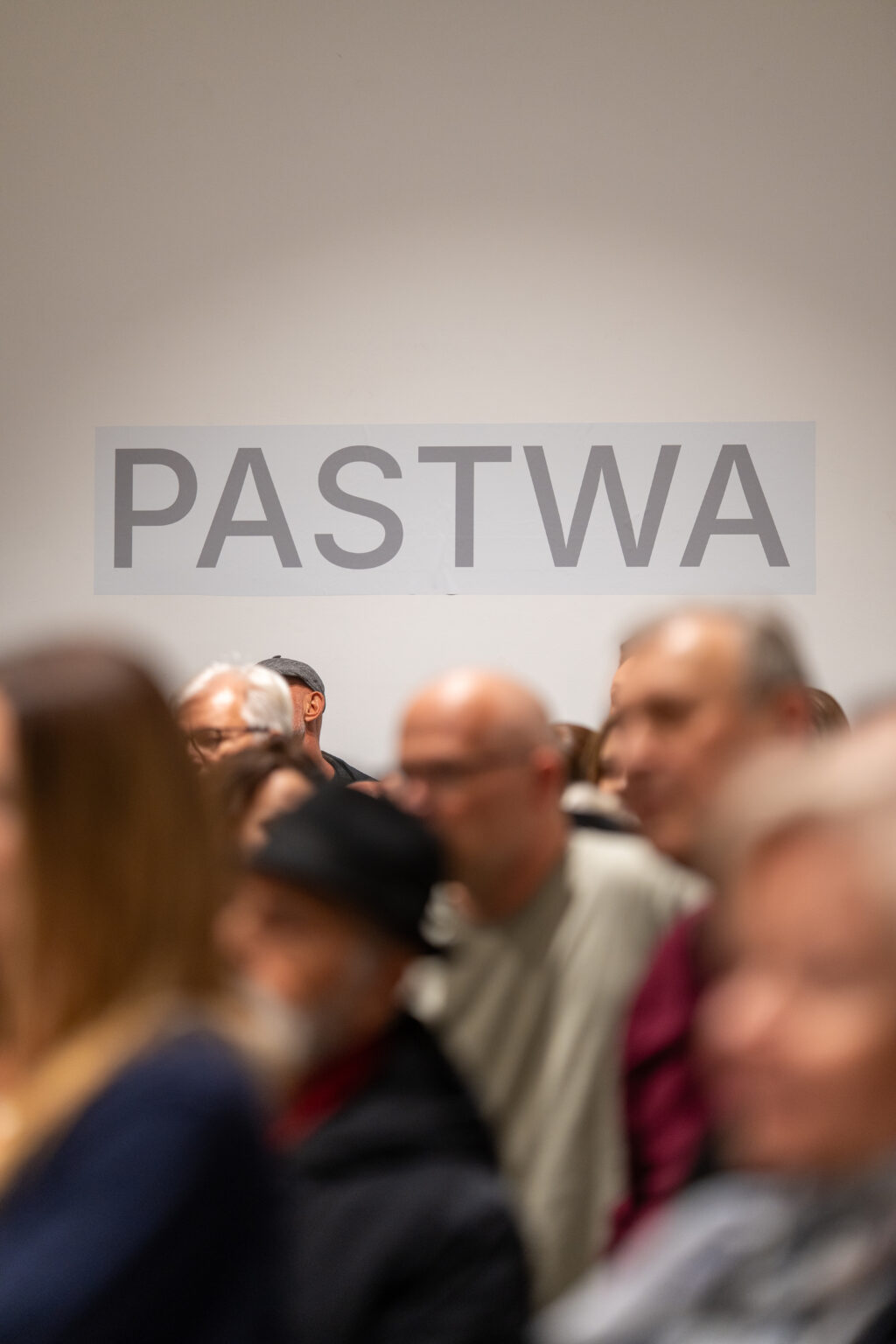 Tłum gości na wystawie „PASTWA” – rozmyte sylwetki na tle napisu.