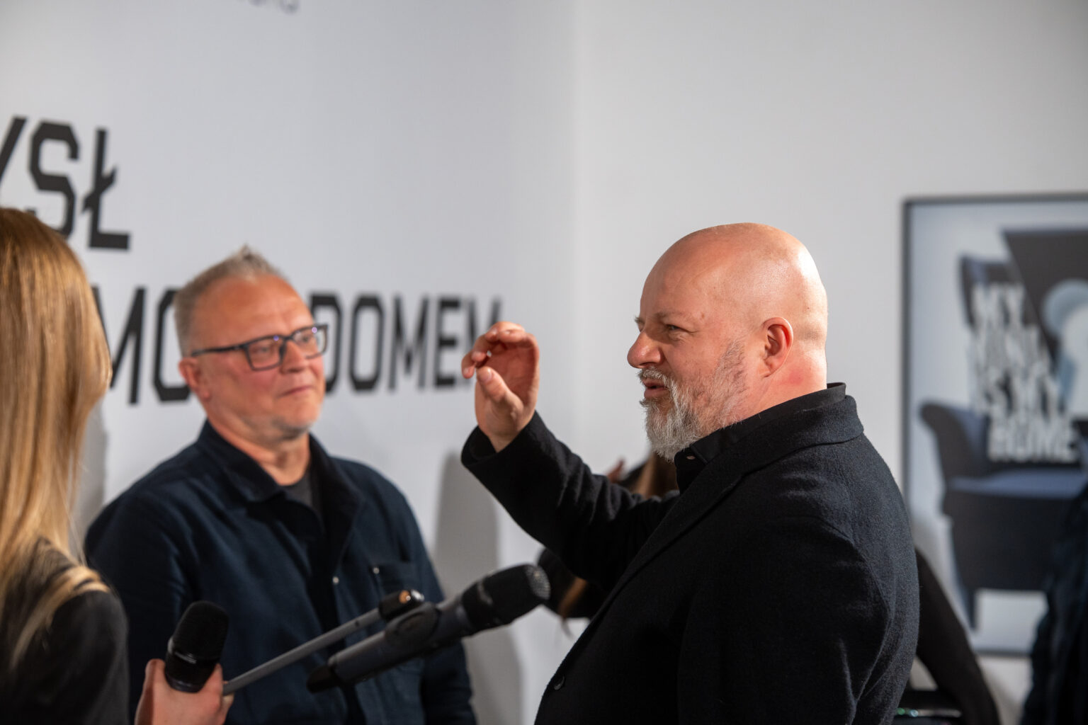 Mężczyzna z brodą udzielający wywiadu do mikrofonów w galerii sztuki.