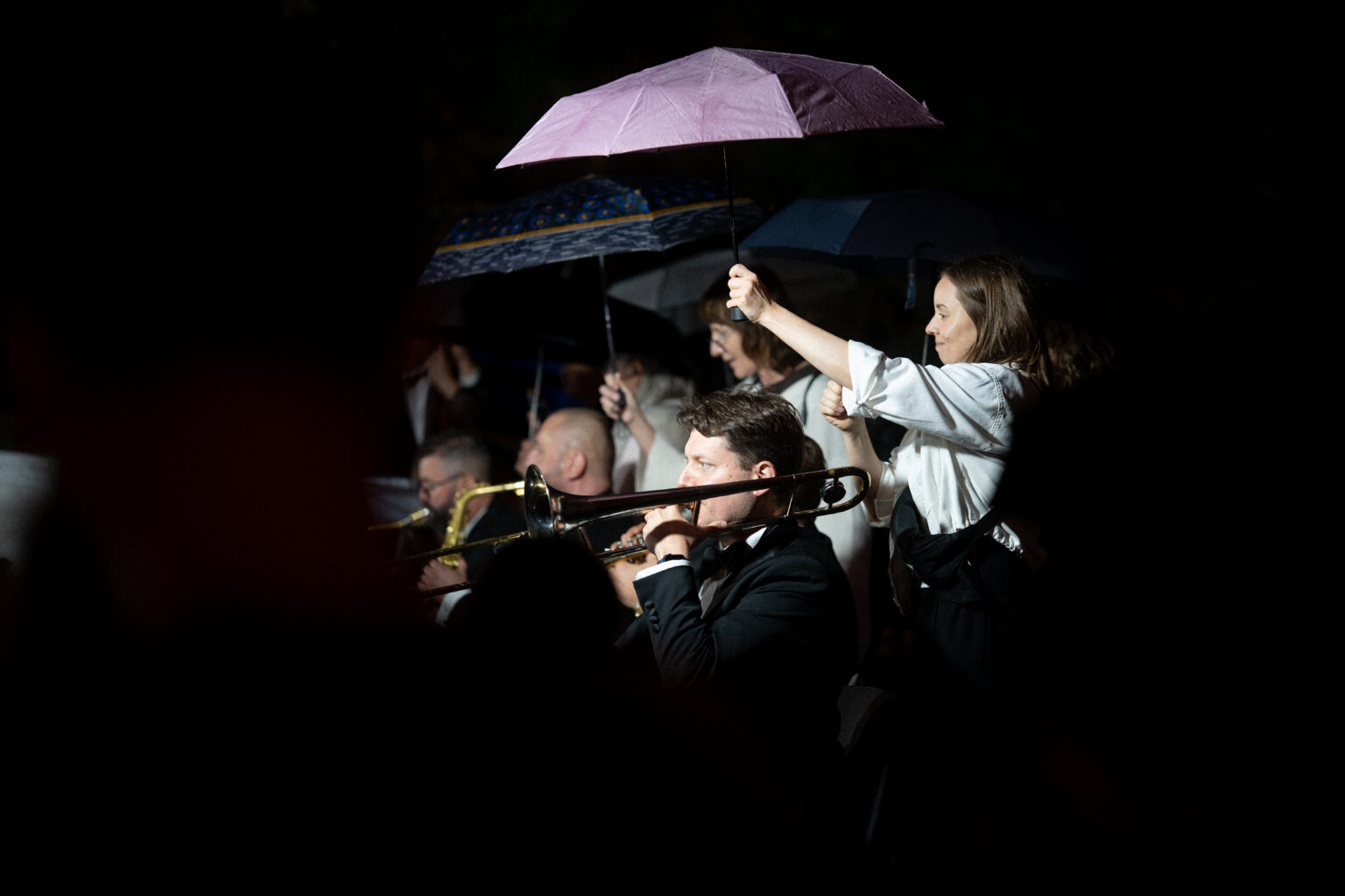 Kobieta trzyma parasol nad puzonistą podczas nocnego koncertu.